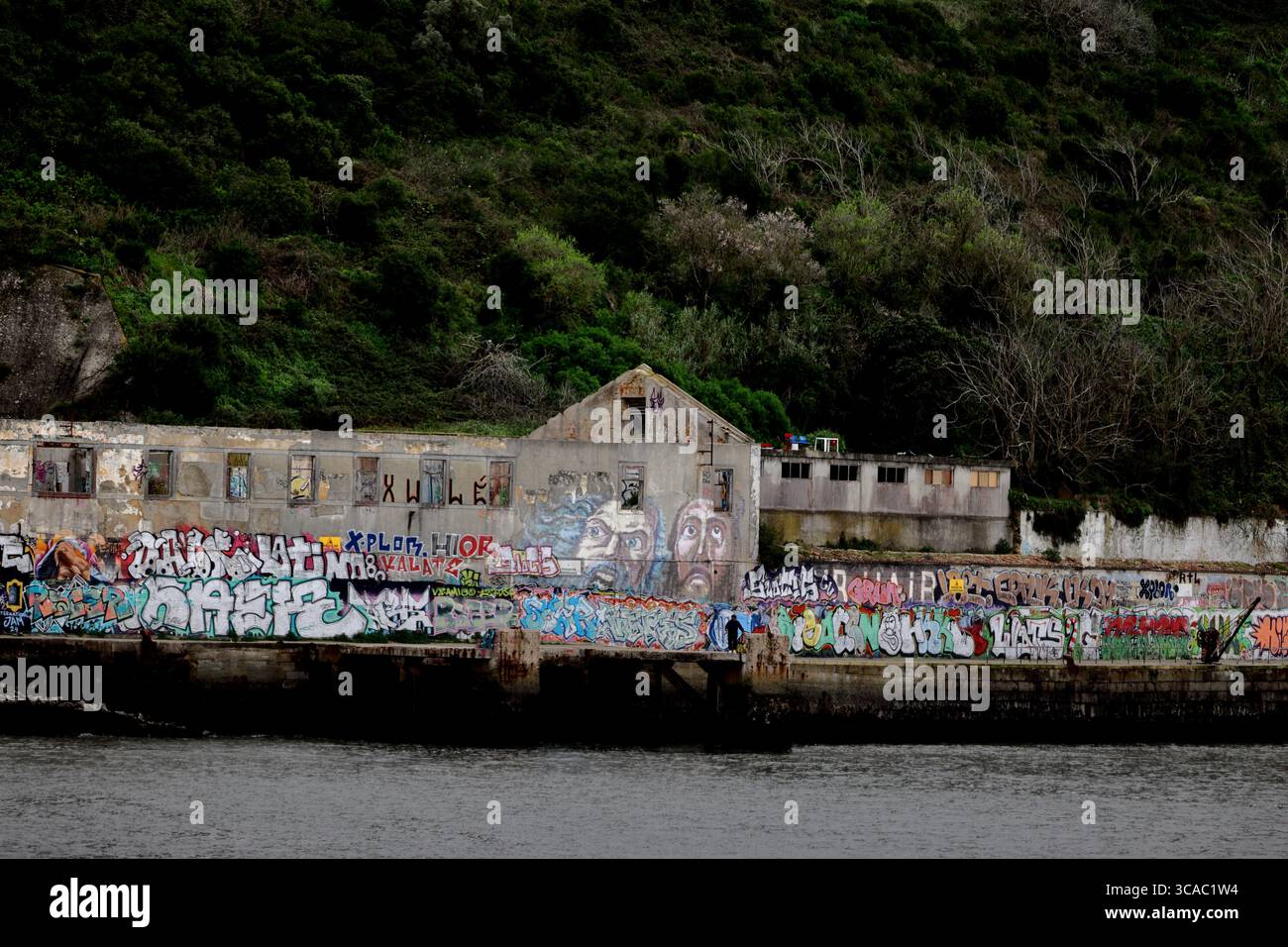 Entrepôts riverains recouverts de graffitis à Praia das Lavadeiras, Almada, Lisbonne, au-dessous d'une colline verdoyante, capturant le caractère côtier urbain graveleux. Banque D'Images