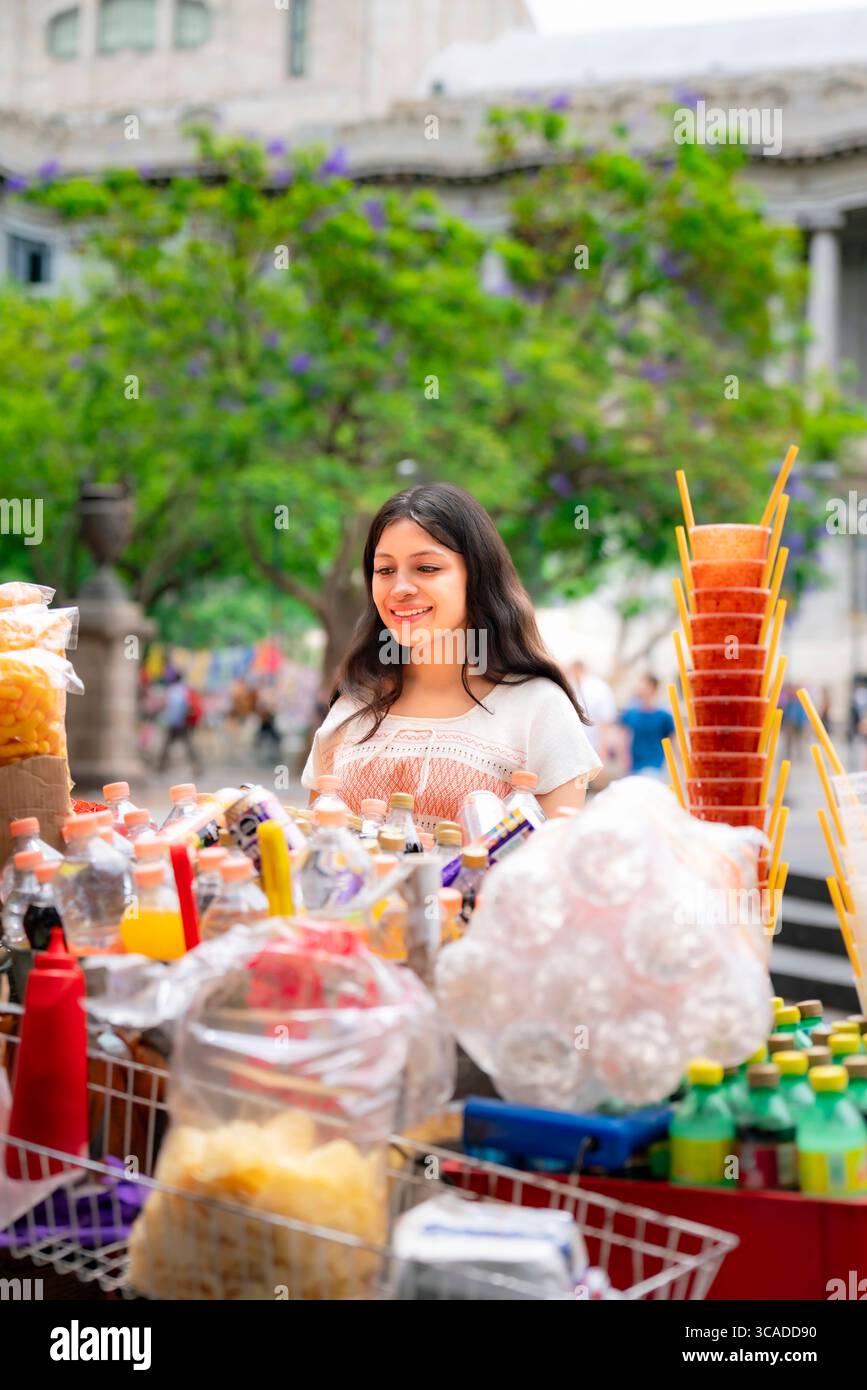 Jeune Latina souriante dans une robe blanche et corail explore un stand de nourriture de rue dynamique plein de frites, de boissons et de bonbons à Alameda Central, à côté du Banque D'Images