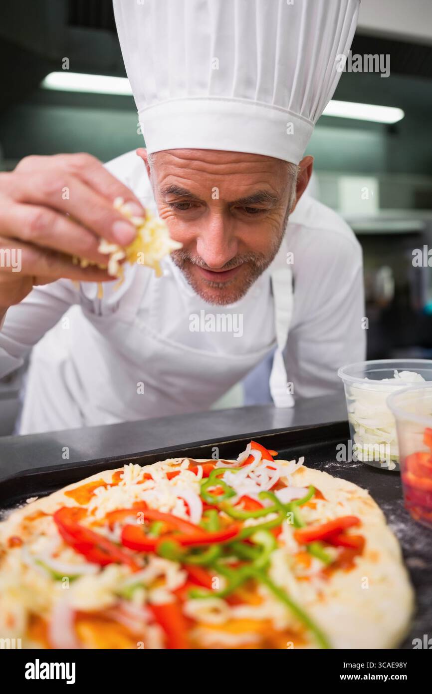 Homme chef portant une veste toque blanche saupoudrant du fromage râpé sur la croûte de pizza au comptoir de la cuisine Banque D'Images