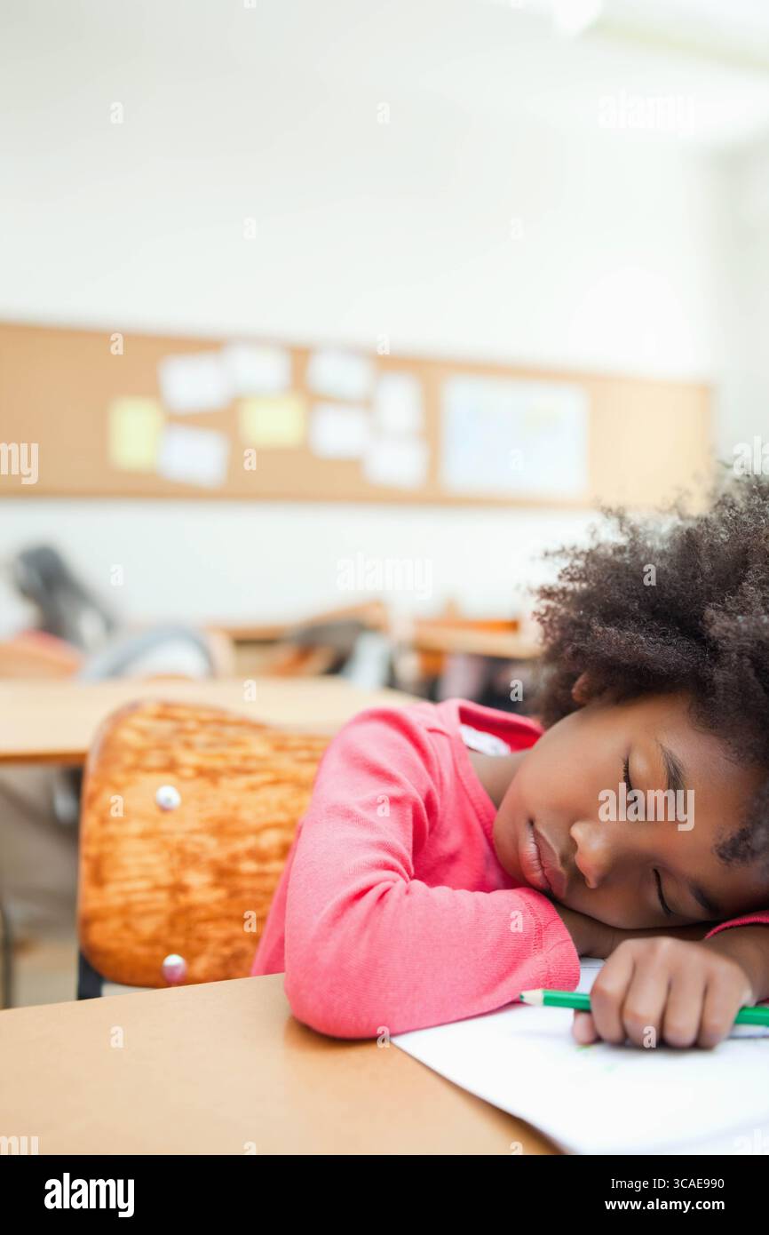 Fille afro-américaine reposant la tête sur les bras au bureau de la classe avec crayon vert, papier, espace de copie Banque D'Images