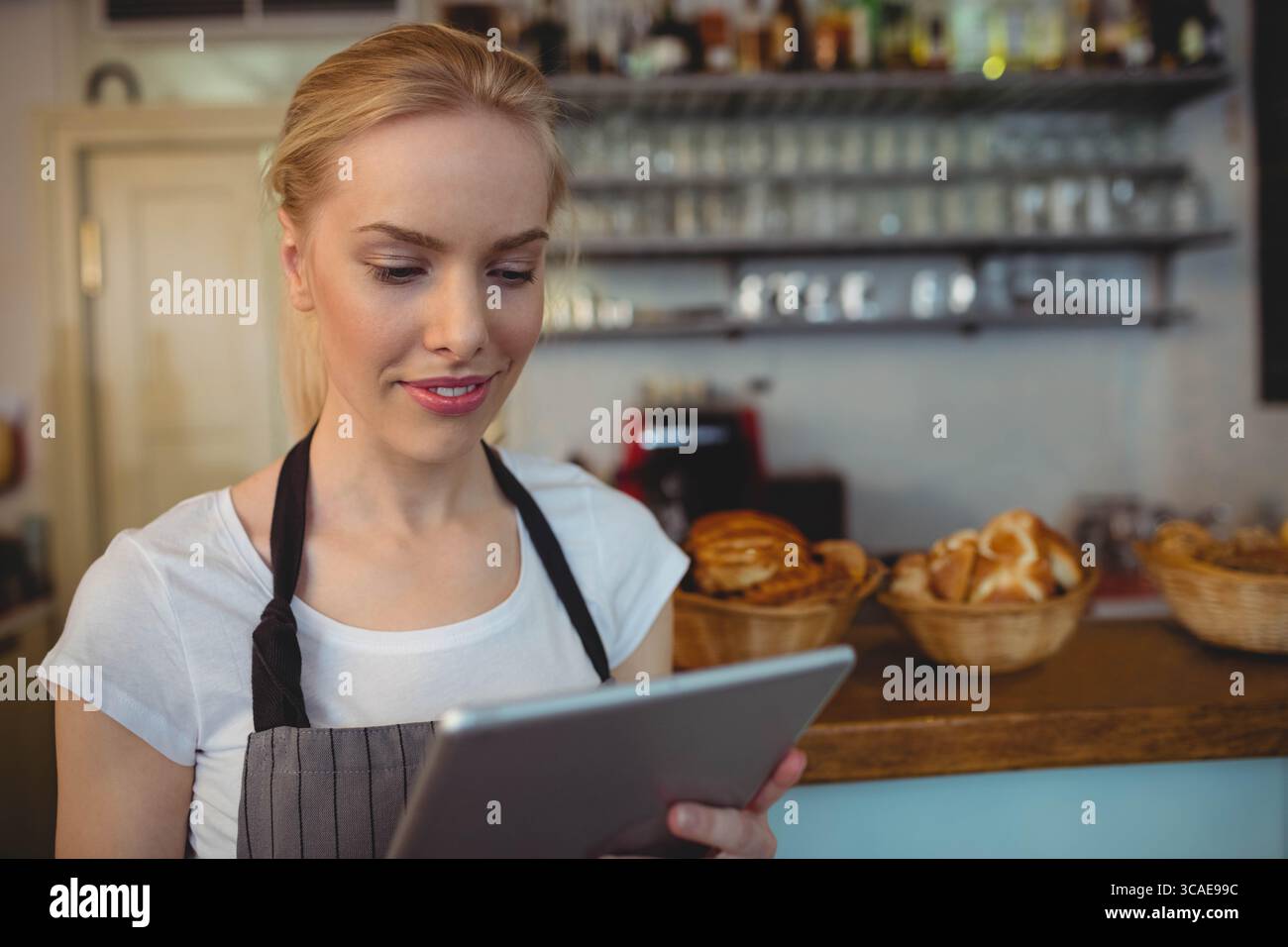 Femme portant un tablier rayé vérifiant l'écran de tablette au comptoir de café avec des pâtisseries et une machine à café Banque D'Images