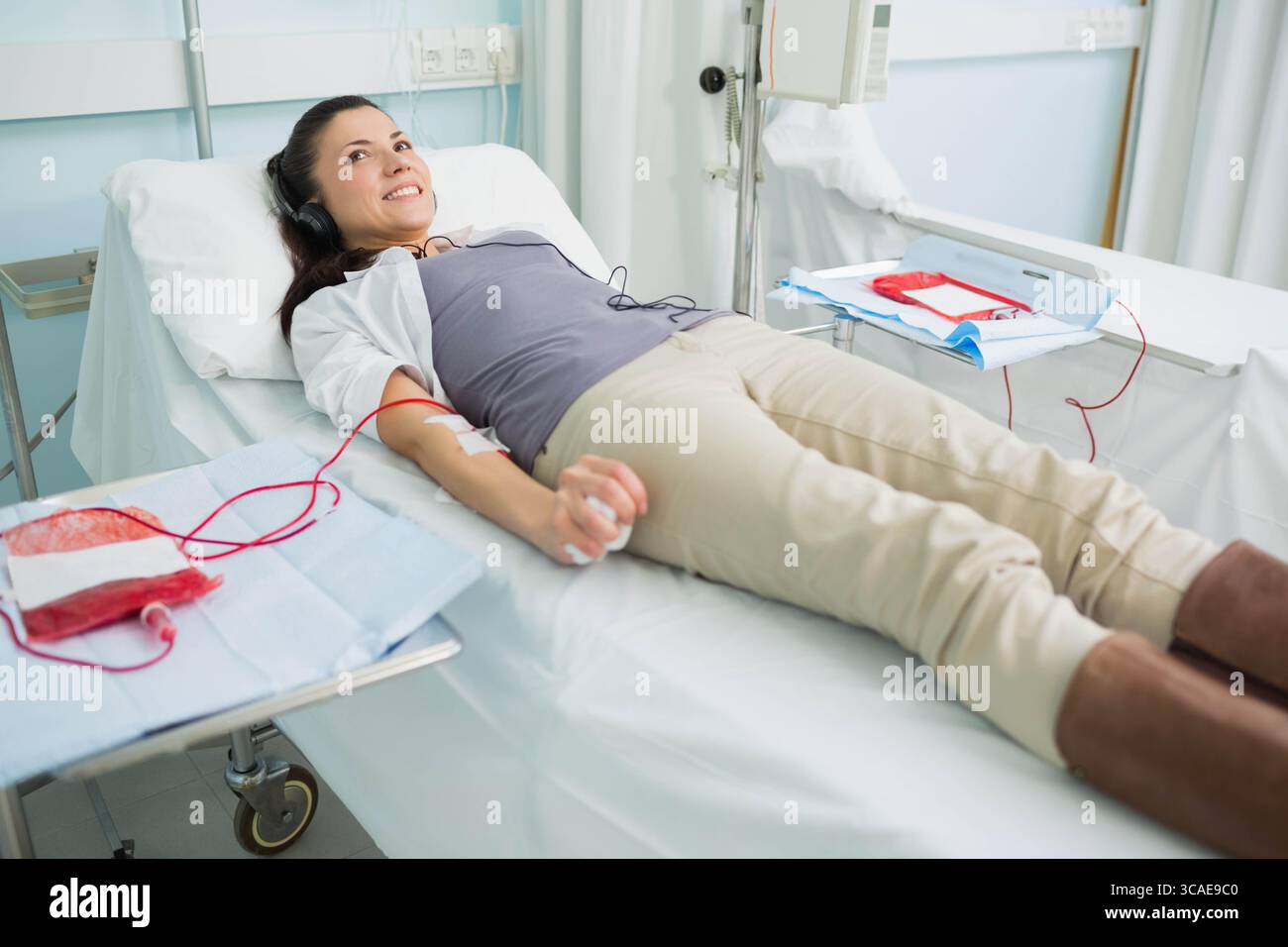 Donneuse couchée sur le lit d'hôpital avec poche de sang, aiguille et écouteurs dans la salle de don Banque D'Images