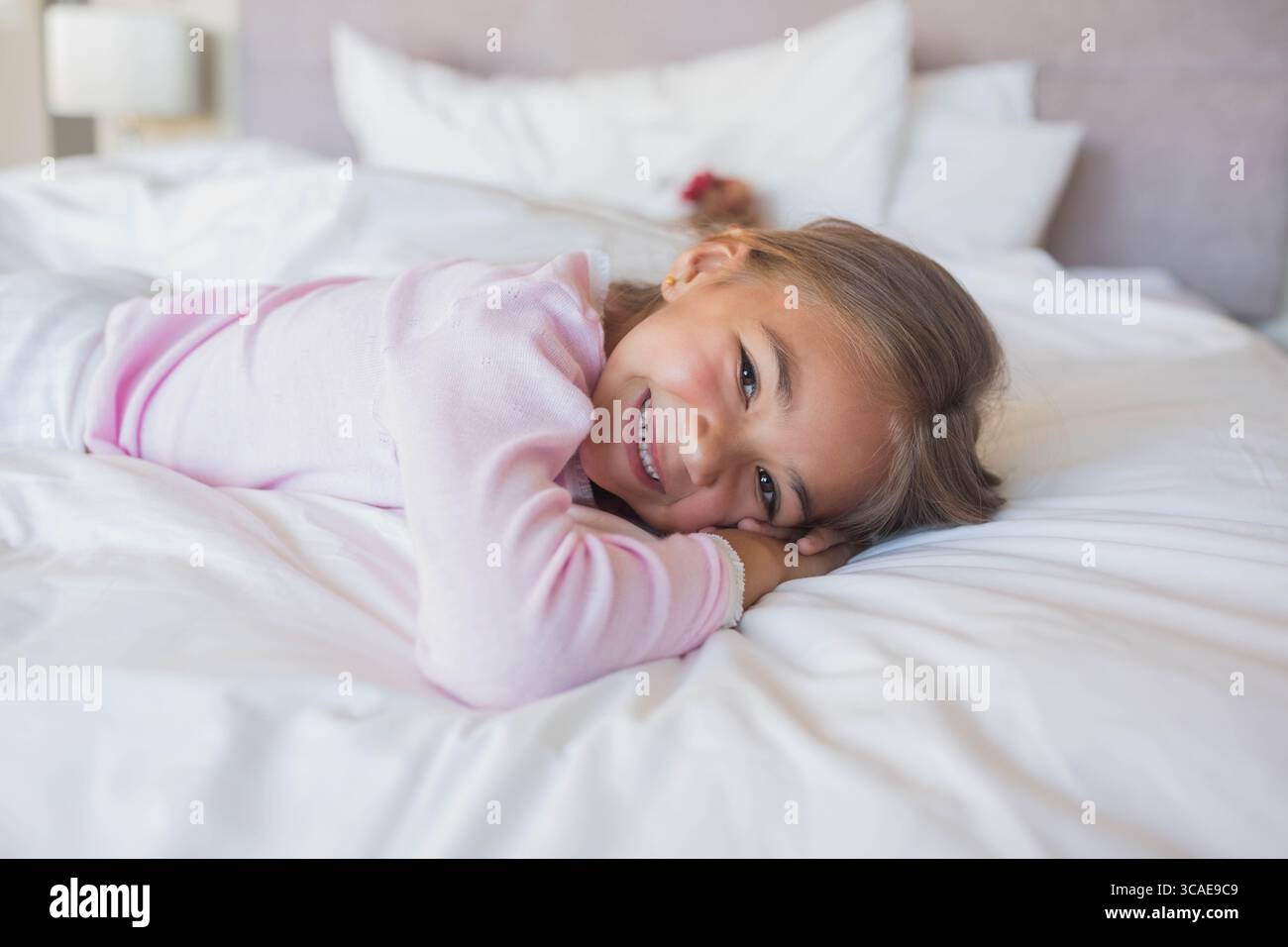Fille couchée sur le ventre sur des draps blancs dans la chambre, soutenant la tête sur les mains, souriant à côté de la lampe Banque D'Images