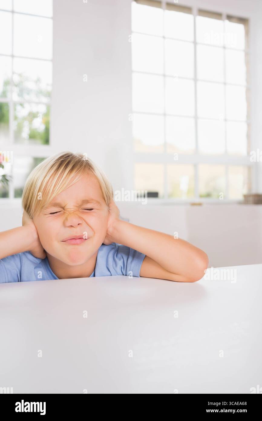 Garçon pressant les mains sur les oreilles et plissant au dessus de la table blanche à l'intérieur de la pièce près de la fenêtre à motif quadrillé Banque D'Images