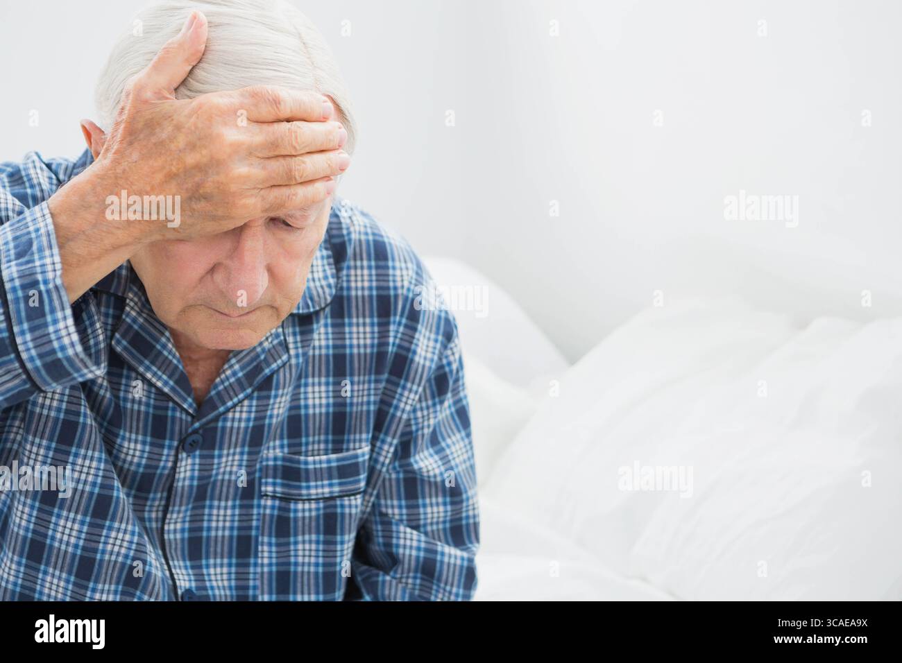 Homme âgé assis sur le lit portant un pyjama à carreaux tenant la main au front sur des oreillers dans la chambre Banque D'Images