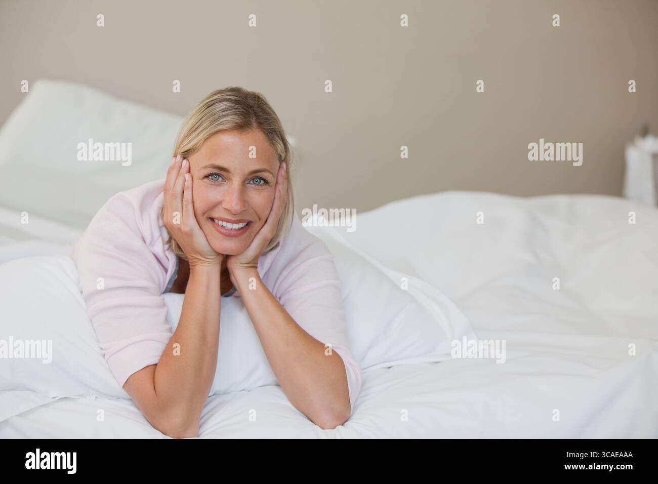 Femme adulte souriante couchée sur une literie blanche portant un oreiller enveloppant rose clair dans la chambre Banque D'Images