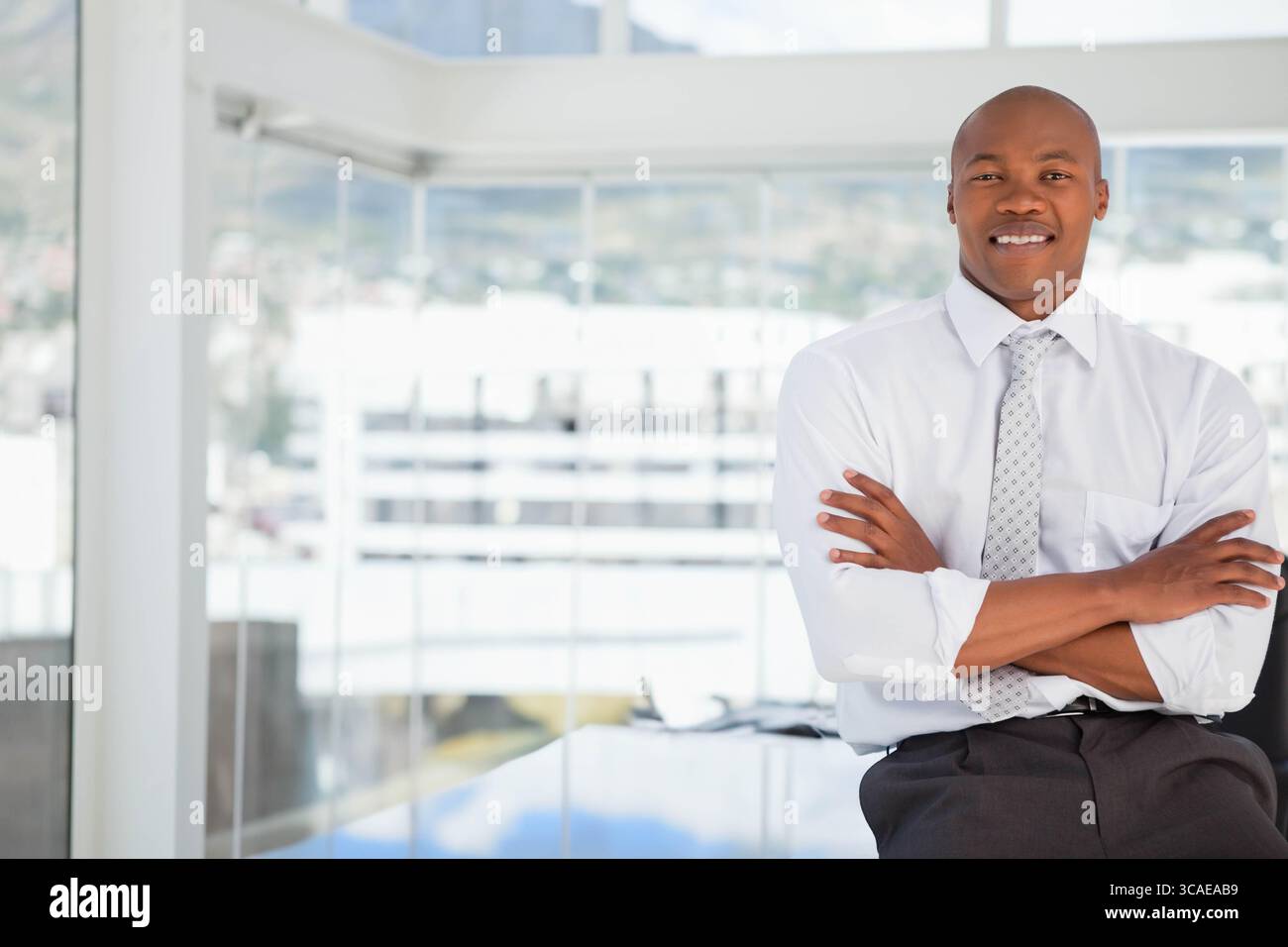 Homme d'affaires afro-américain appuyé contre le bureau par les fenêtres du sol au plafond du bureau, espace de copie Banque D'Images
