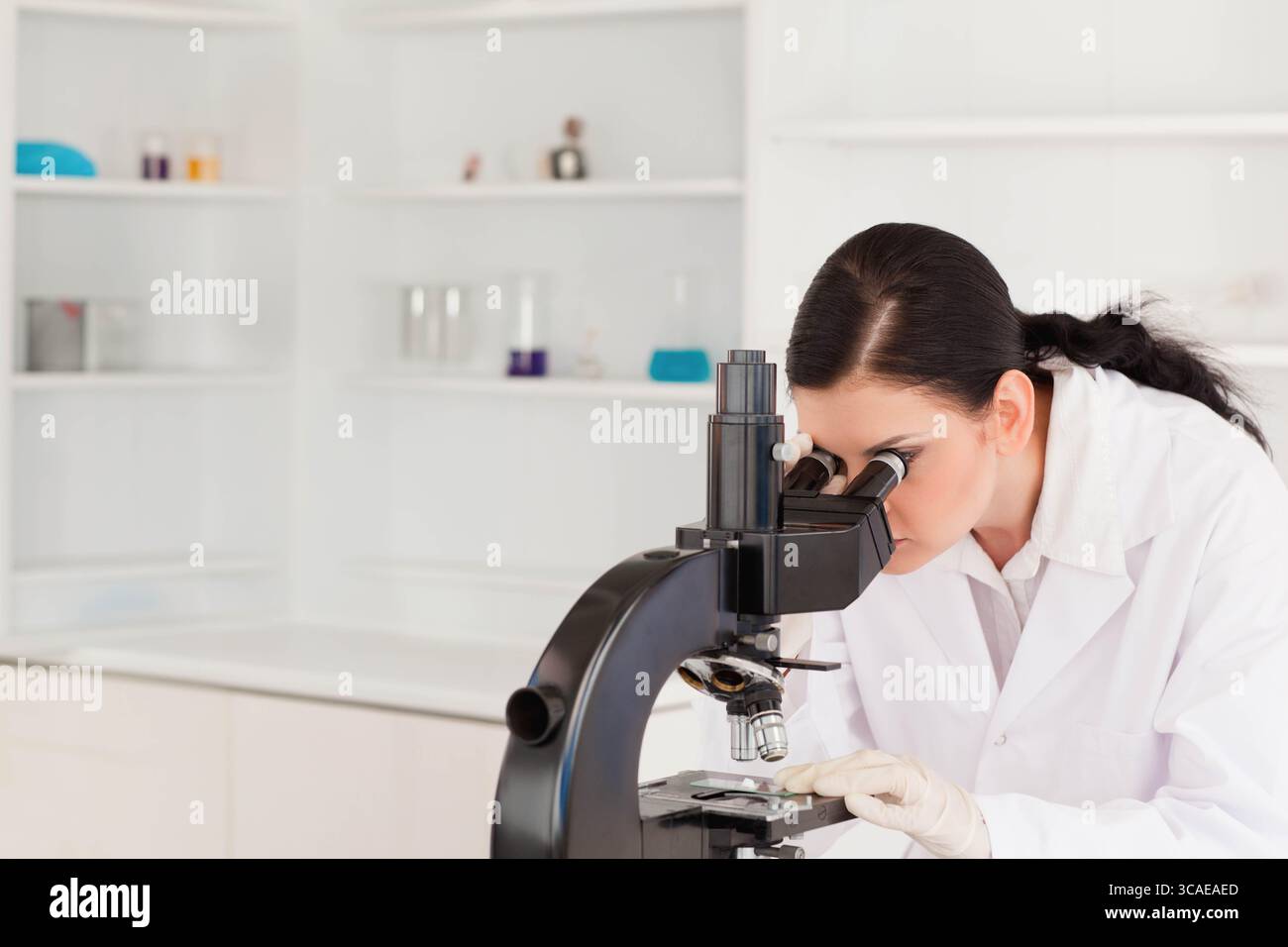 Femme en blouse de laboratoire et gants regardant dans le microscope sur le banc de laboratoire avec la verrerie, copier l'espace Banque D'Images