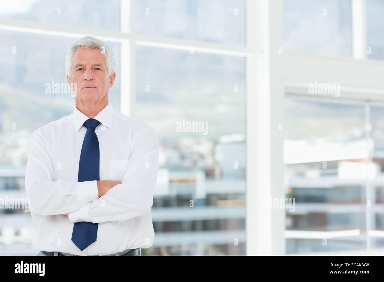 Homme senior debout devant de grandes fenêtres en verre dans le bureau portant une chemise blanche cravate marine, espace de copie Banque D'Images