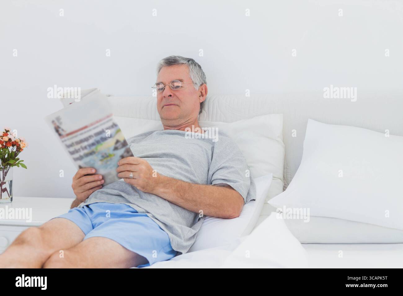Homme senior incliné contre la tête de lit lisant le journal sur le lit près de la table de nuit avec des fleurs oranges Banque D'Images
