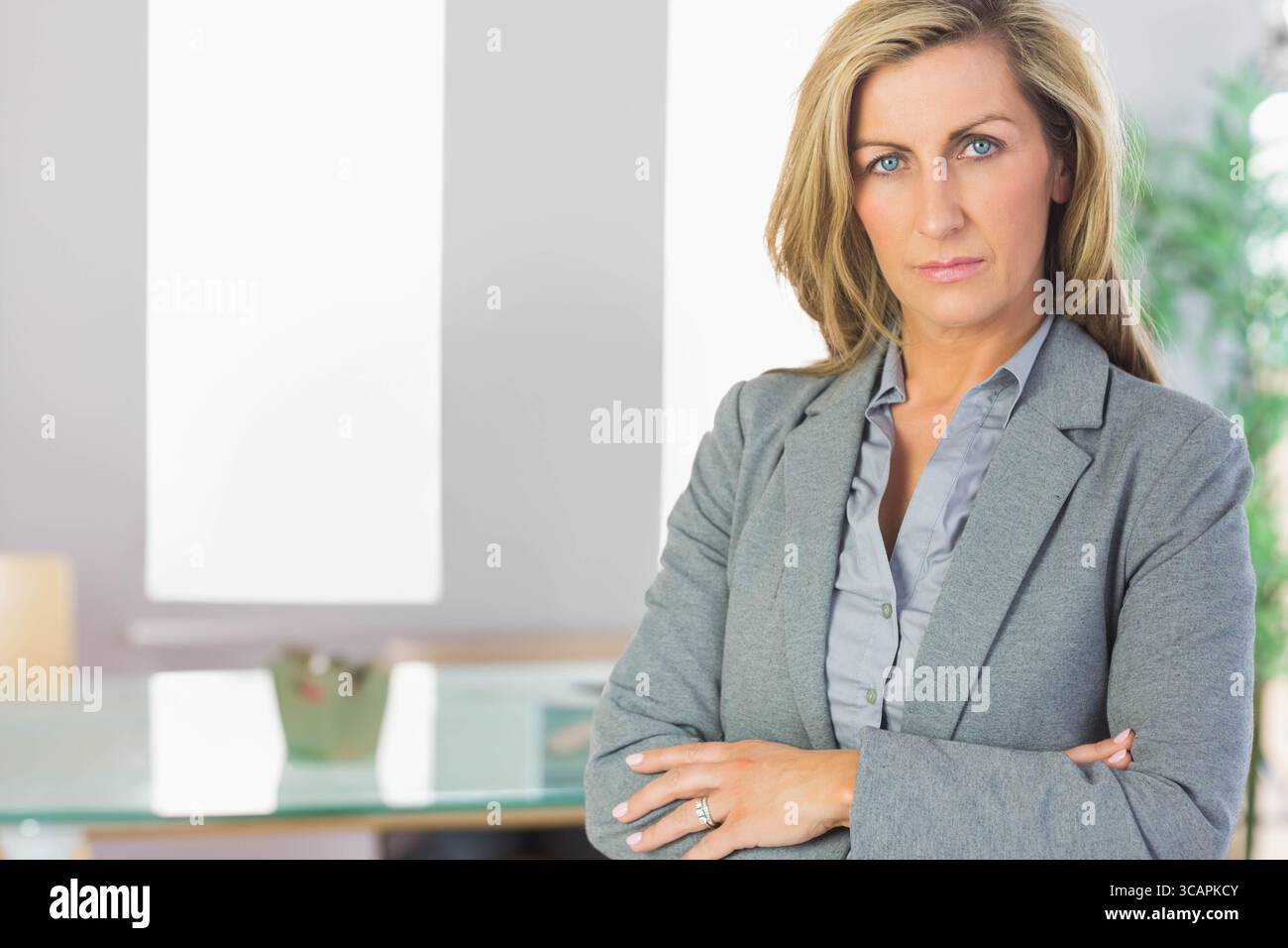 Femme adulte moyenne debout bras croisés dans le bureau avec bureau en verre, panneau blanc, espace de copie Banque D'Images