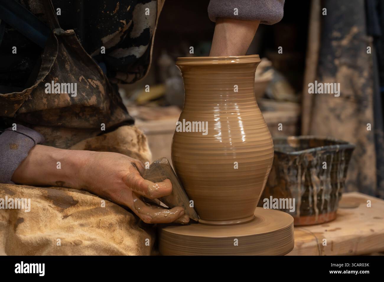 Gros plan des mains d’un potier recouvertes d’argile humide alors qu’elles façonnent soigneusement un vase en céramique sur une roue de poterie. L'artisan utilise un outil en bois pour affiner t Banque D'Images