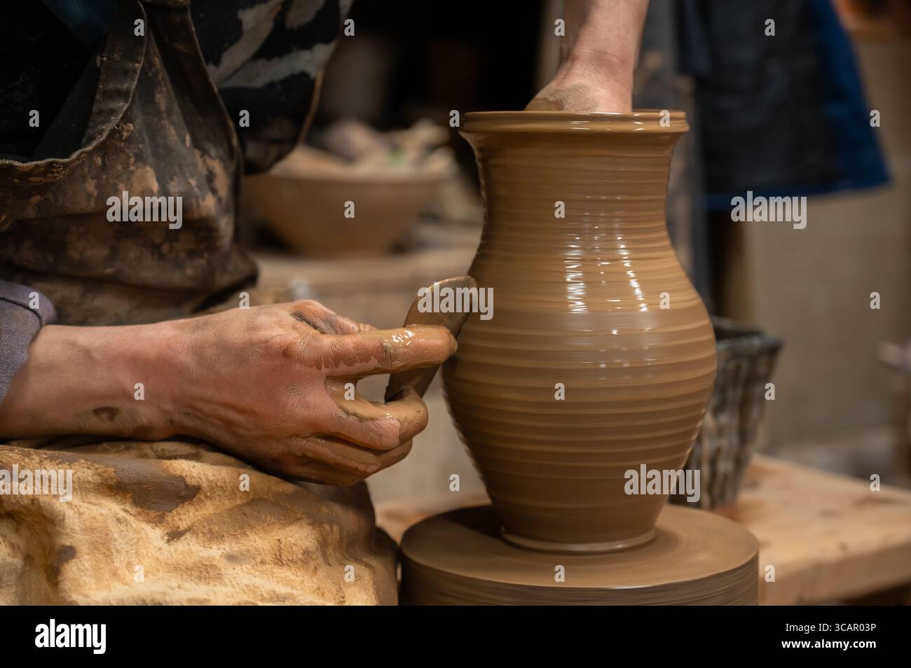 Gros plan des mains d’un potier recouvertes d’argile humide alors qu’elles façonnent soigneusement un vase en céramique sur une roue de poterie. L'artisan utilise un outil en bois pour affiner t Banque D'Images