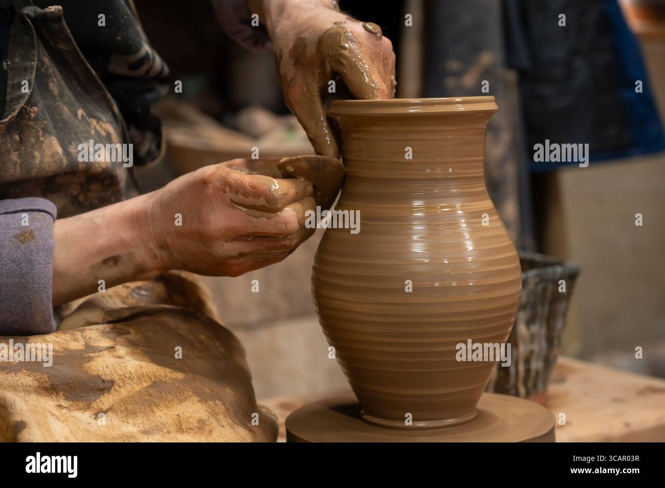 Gros plan des mains d’un potier recouvertes d’argile humide alors qu’elles façonnent soigneusement un vase en céramique sur une roue de poterie. L'artisan utilise un outil en bois pour affiner t Banque D'Images
