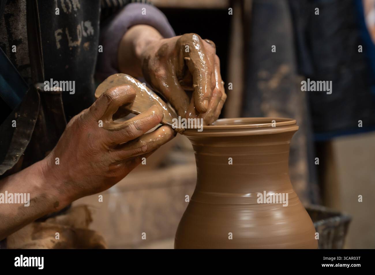 Gros plan des mains d’un potier recouvertes d’argile humide alors qu’elles façonnent soigneusement un vase en céramique sur une roue de poterie. L'artisan utilise un outil en bois pour affiner t Banque D'Images