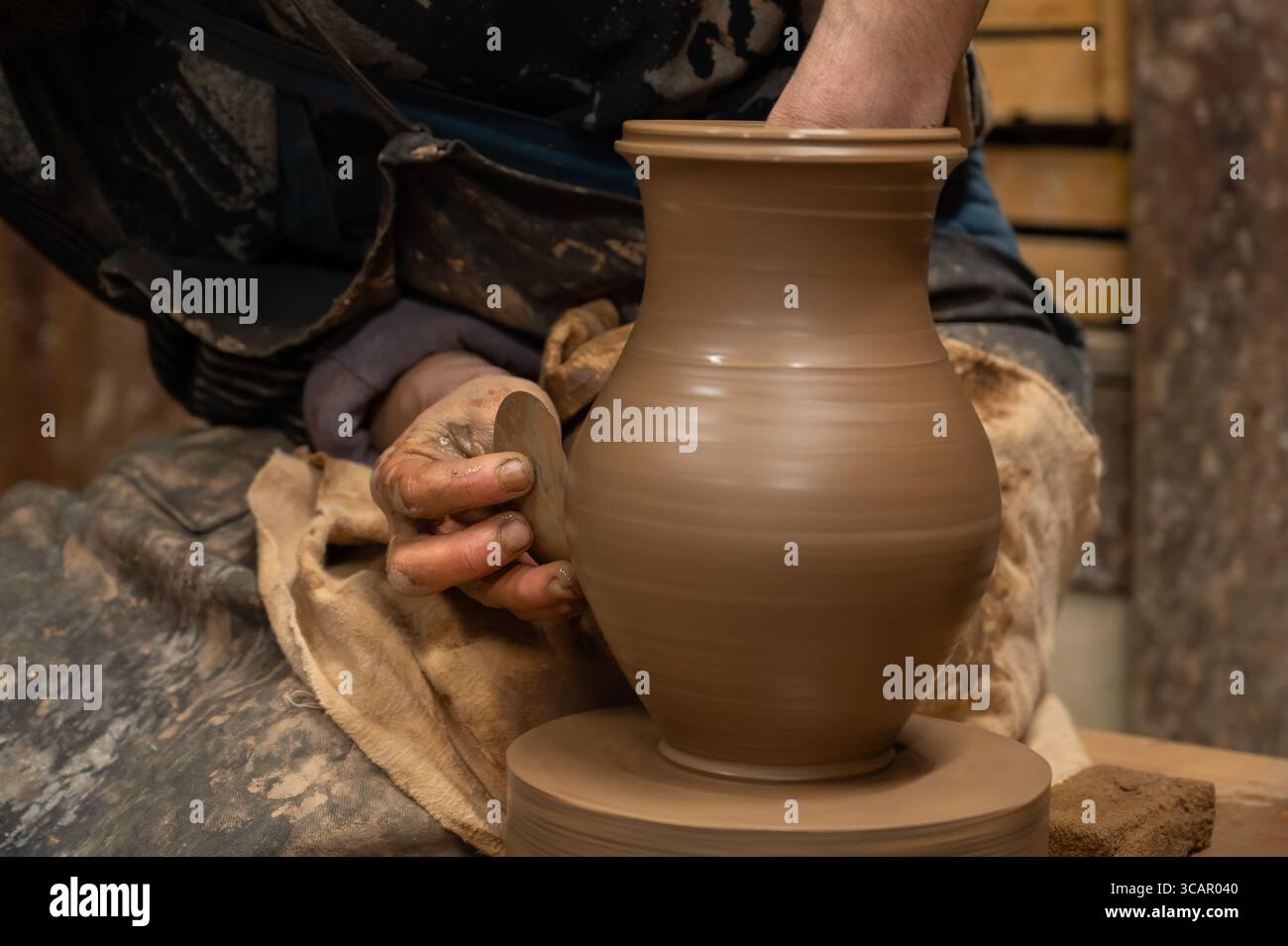 Gros plan des mains d’un potier recouvertes d’argile humide alors qu’elles façonnent soigneusement un vase en céramique sur une roue de poterie. L'artisan utilise un outil en bois pour affiner t Banque D'Images
