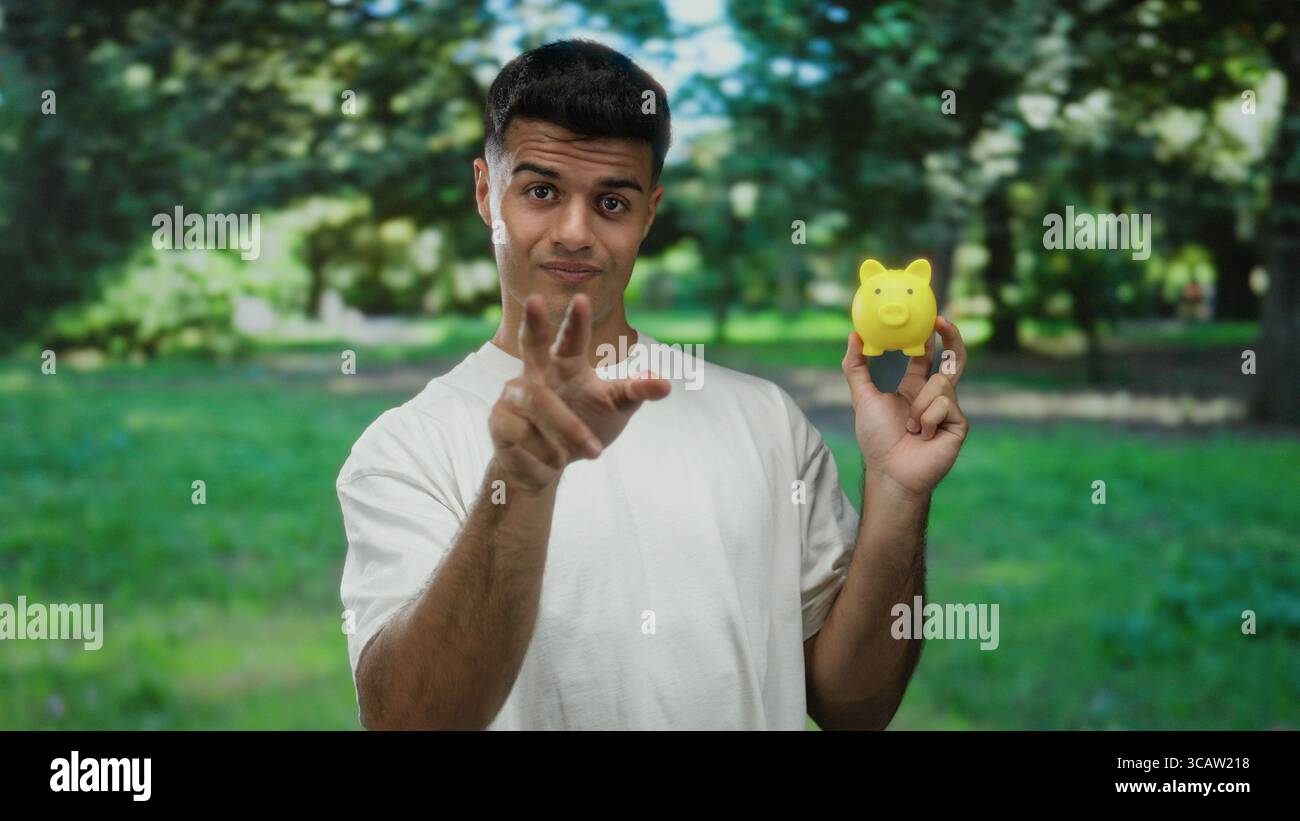 Jeune homme hispanique fait un geste tout en tenant une tirelire jaune dans un cadre de parc extérieur, portant une chemise blanche, transportant un joueur et observ Banque D'Images