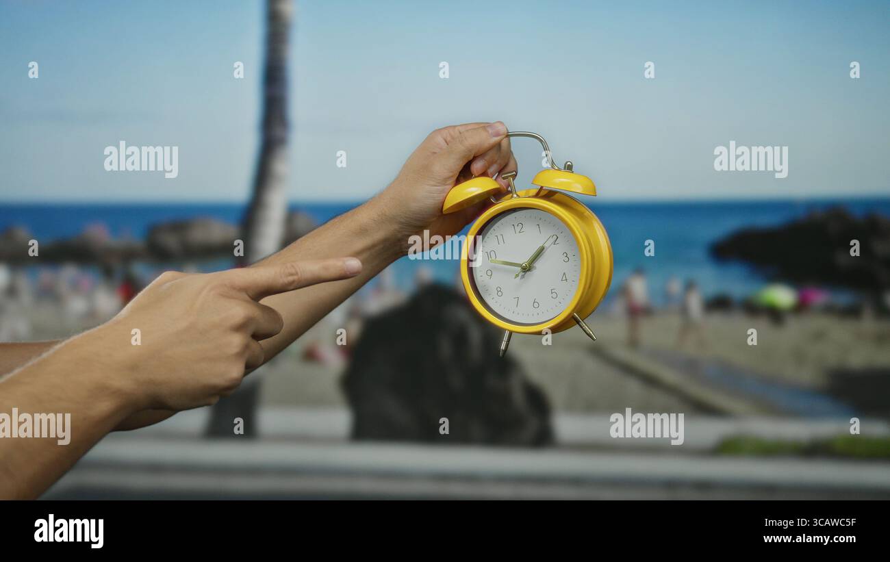 Main masculine tenant un réveil jaune à la plage avec fond flou de bord de mer sous le ciel bleu indiquant le concept de temps dans une atmosphère côtière détendue. Banque D'Images