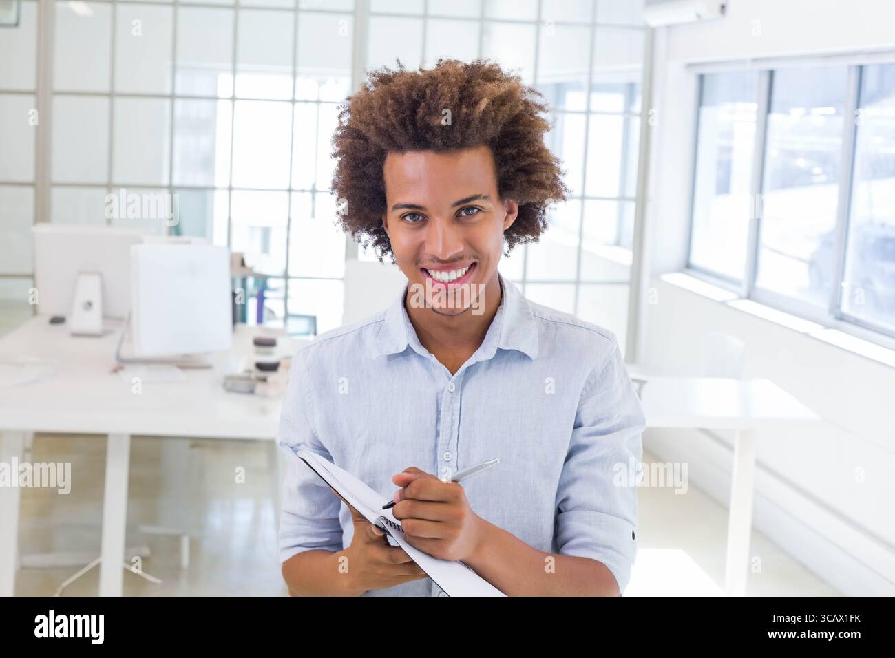 Homme afro-américain portant une chemise bleu clair au bureau dans un bureau ouvert tenant un bloc-notes et un stylo Banque D'Images