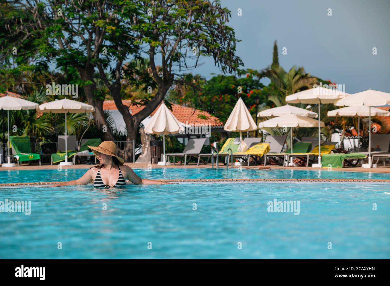 Une femme en bikini noir et blanc est assise dans une piscine. La piscine est entourée de parasols et de chaises Banque D'Images