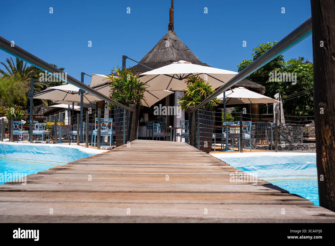 Un pont en bois mène à une piscine avec parasols et chaises. Les parapluies sont blancs et les chaises bleues. La scène est relaxante et accueillante Banque D'Images