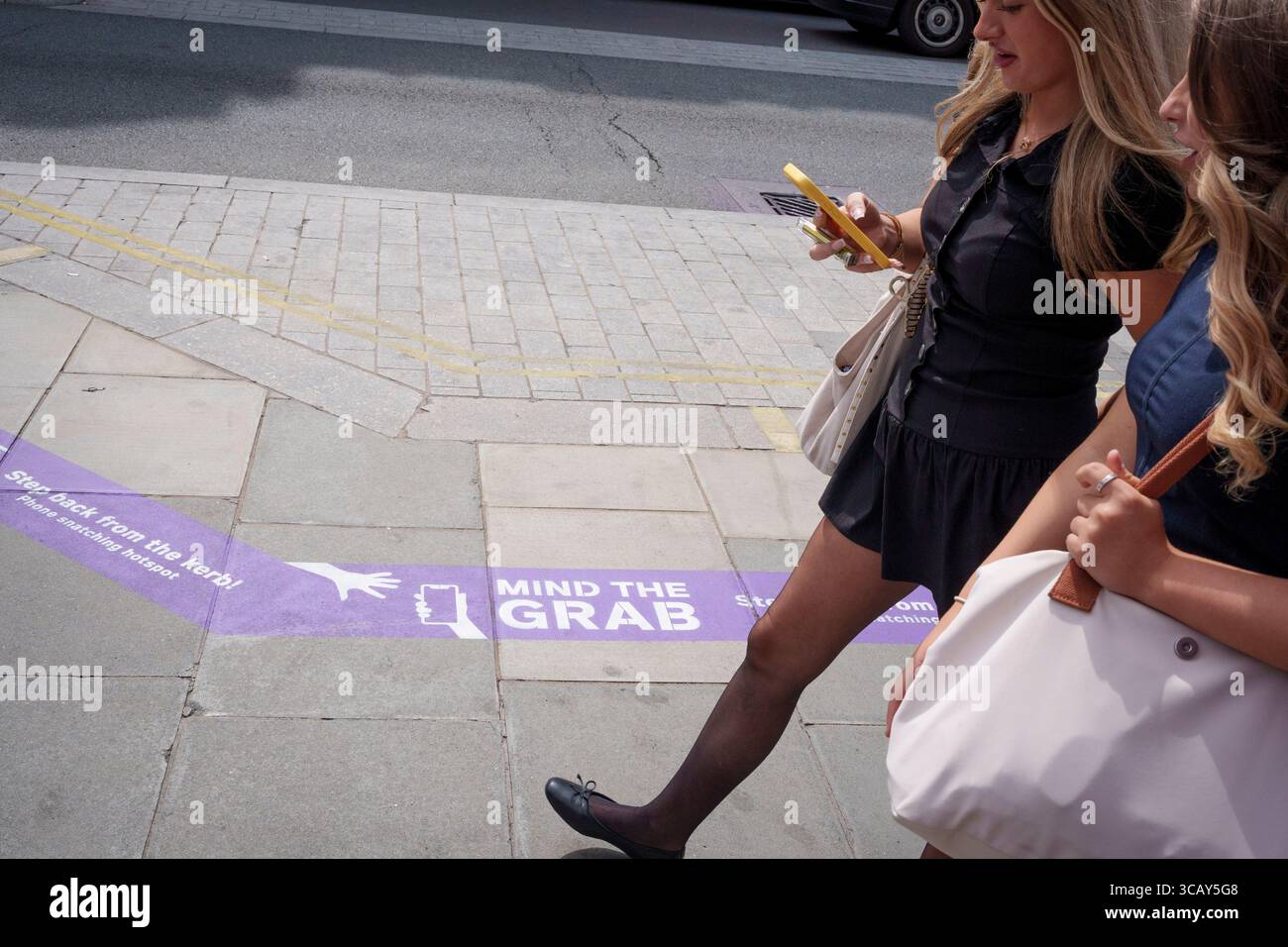 Un utilisateur de téléphone et d'autres piétons passent devant des messages de trottoir sur Oxford Street qui les avertissent de la tendance croissante de la criminalité par téléphone, l'initiative « Mind the Grab » par Westminster, la police met et le détaillant Currys, le 7 août 2025, à Londres, en Angleterre. Les statistiques révèlent qu'en 2024, un téléphone a été volé toutes les 15 minutes à Westminster - 94 par jour et plus de 34 000 au total. Typiquement, ces vols impliquent des criminels sur des cyclomoteurs ou des vélos électriques arrachant des téléphones directement des mains des gens sur des trottoirs fréquentés. Banque D'Images