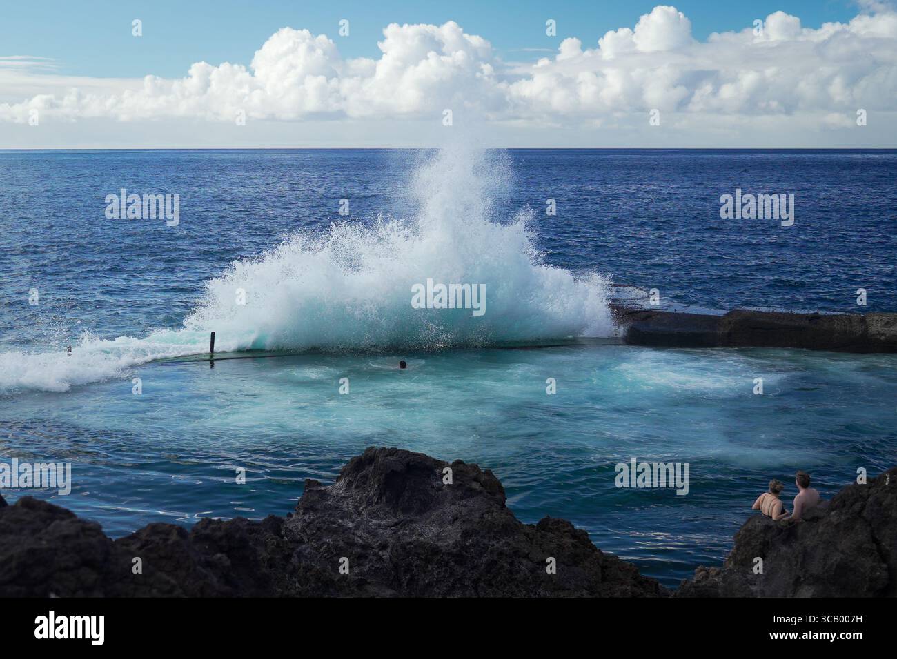 Une puissante vague océanique s'écrase dans une piscine rocheuse naturelle par une journée ensoleillée, tandis que les gens nagent et se détendent près des roches volcaniques sous un ciel bleu avec des nuages moelleux Banque D'Images