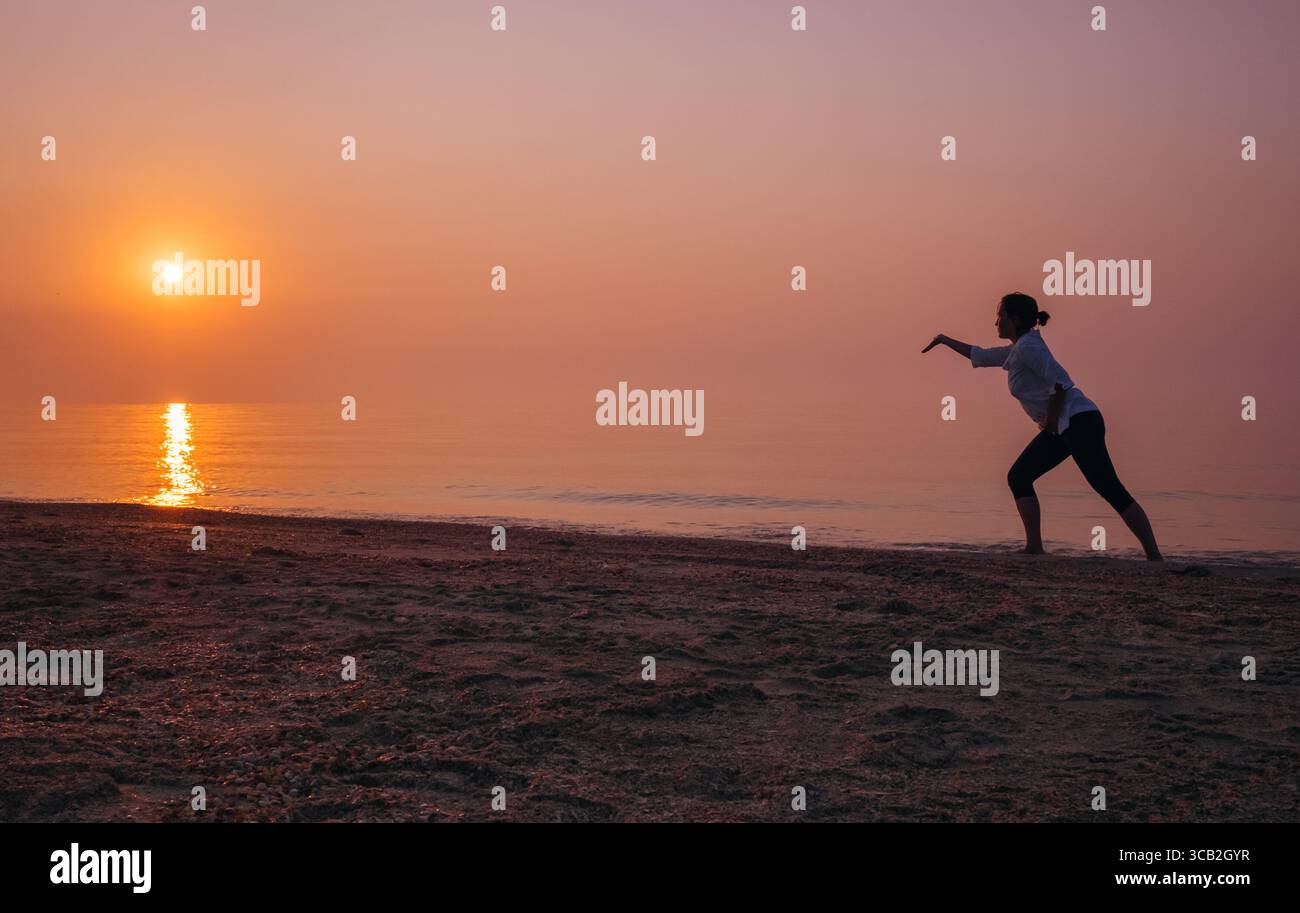 Femme pratiquant le Tai Chi sur la plage au lever du soleil, silhouette gracieuse contre mer calme et ciel orange-rose. Pleine conscience, style de vie bien-être, balance énergétique Banque D'Images