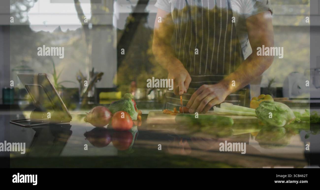 Hacher cuisinier à la maison portant un tablier rayé tranchant des carottes sur l'îlot de cuisine à la maison, avec support de tablette Banque D'Images