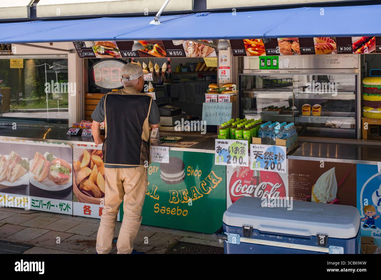 Un homme en vêtements décontractés se tient à un stand de nourriture en plein air, choisissant parmi un menu de collations et de boissons. Le stand de nourriture dispose d'une variété de fast-food Banque D'Images