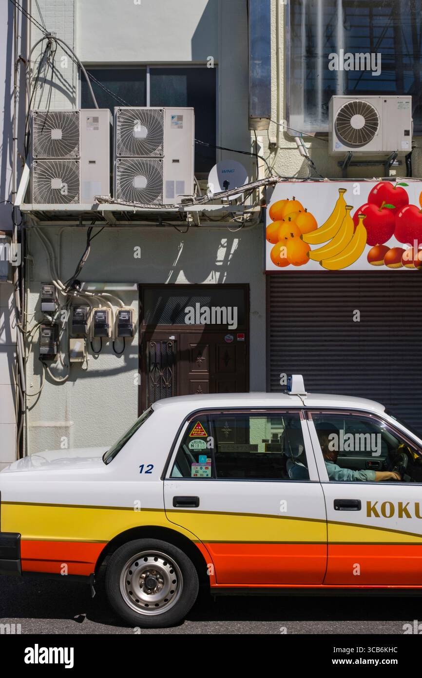Un taxi urbain est garé devant un bâtiment avec des climatiseurs visibles et un panneau de fruits. La scène capture la vie urbaine, Sasebo, Japon Banque D'Images