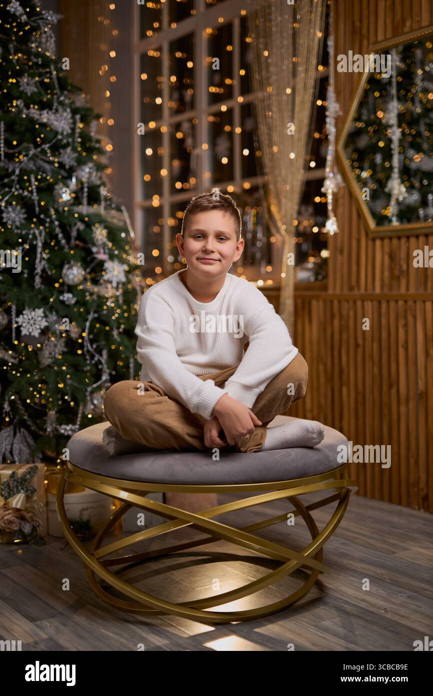 Un garçon souriant dans un pull blanc est assis avec soin pour une photo de vacances, avec un arbre de Noël décoré et des lumières scintillantes en arrière-plan Banque D'Images