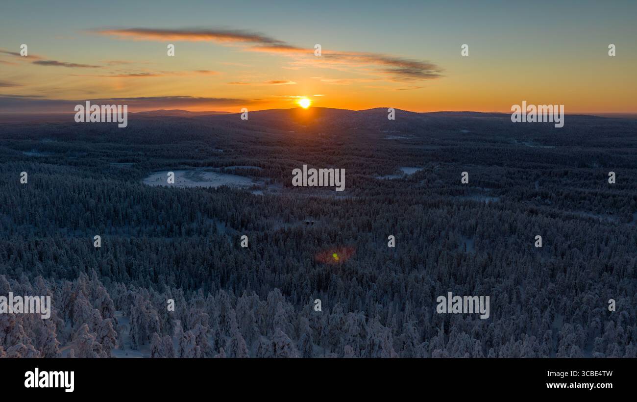 Vue aérienne du soleil se levant sur les forêts enneigées et les collines, projetant une lueur dorée à travers le paysage hivernal, Salla, Laponie, Finlande. Banque D'Images
