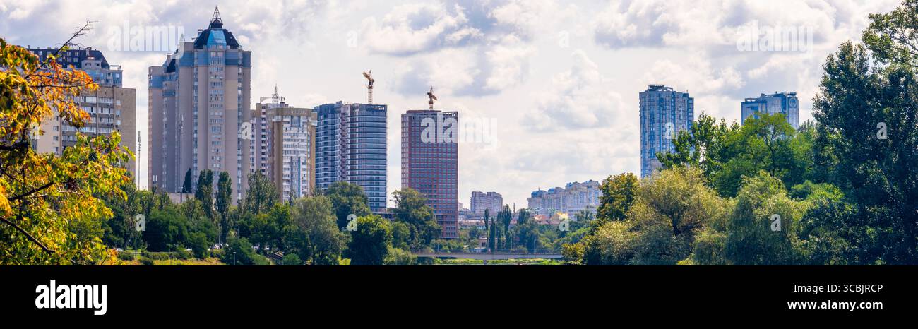 Des arbres verts luxuriants encadrent les bâtiments résidentiels modernes et les grues de construction sous un ciel nuageux d'été à Rusanivka, Kiev, avec la channe de la rivière Dnipro Banque D'Images
