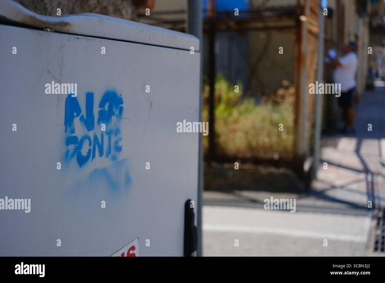 Messine, Sicile, Italie – août 2025. Le vice-premier ministre et ministre de l’infrastructure, Matteo Salvini, assiste à un déjeuner au restaurant Grecale pour célébrer l’approbation par le CIPESS du projet controversé du pont du détroit de Messine. Sur la plage voisine, les baigneurs partagent le sable avec les manifestants tenant un drapeau « No Ponte » (No Bridge) contre la construction. Dans l'eau, un bateau de pêche traditionnel de Messine appelé felouca, utilisé pour la chasse à l'espadon, est décoré de drapeaux italiens de la Lega Nord de droite. Villa San Giovanni en Calabre est visible en arrière-plan. Banque D'Images
