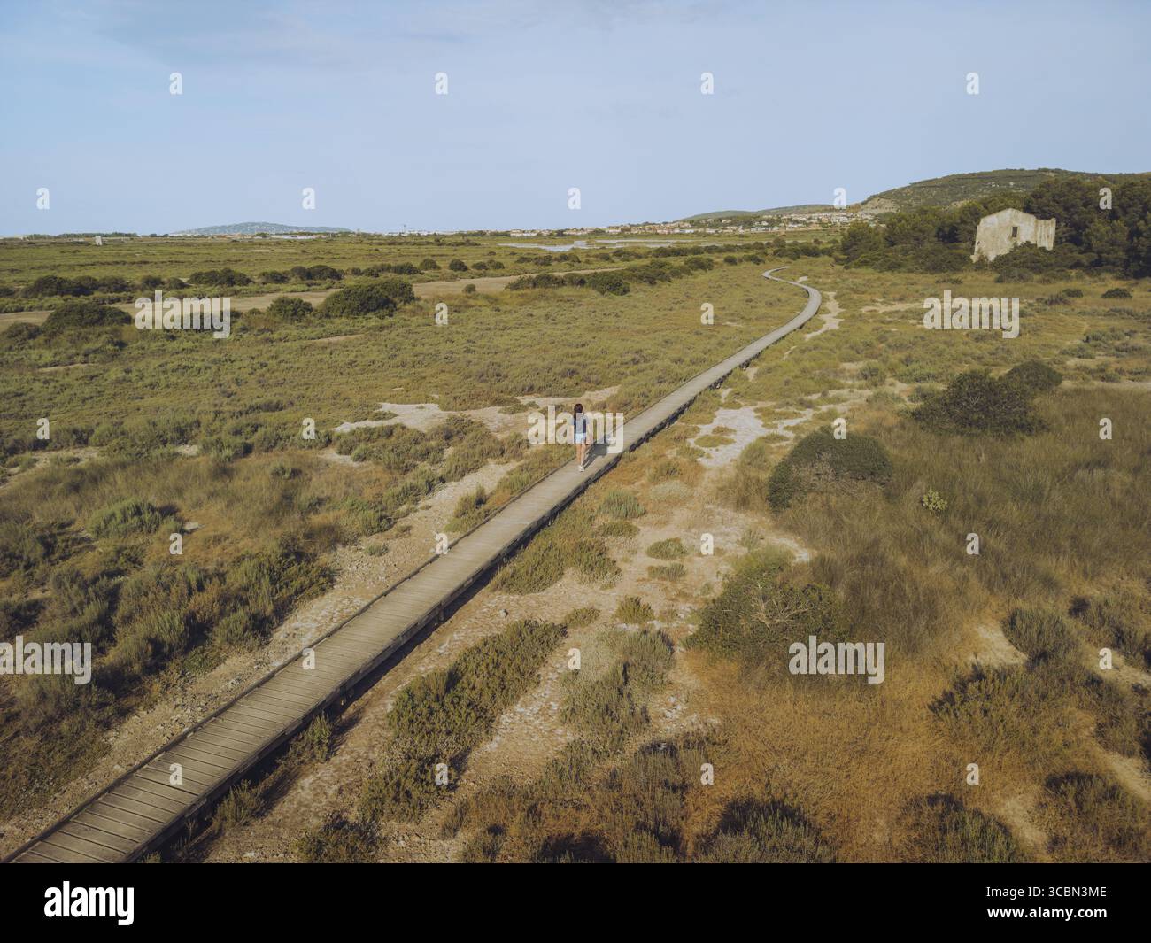 Vue aérienne d'une figure solitaire se promenant le long d'un chemin en bois à travers un paysage de teintes ocre et verdoyantes vers la chaîne de montagnes lointaine de Gardiole, Frontignan, Occitanie, France. Banque D'Images