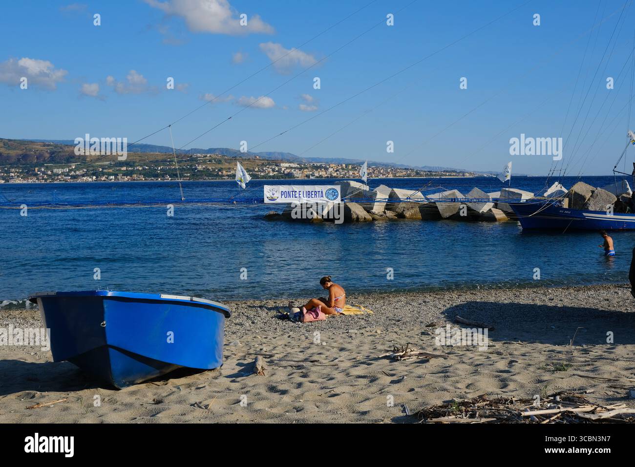 Messine, Sicile, Italie – août 2025. Le vice-premier ministre et ministre de l’infrastructure, Matteo Salvini, assiste à un déjeuner au restaurant Grecale pour célébrer l’approbation par le CIPESS du projet controversé du pont du détroit de Messine. Sur la plage voisine, les baigneurs partagent le sable avec les manifestants tenant un drapeau « No Ponte » (No Bridge) contre la construction. Dans l'eau, un bateau de pêche traditionnel de Messine appelé felouca, utilisé pour la chasse à l'espadon, est décoré de drapeaux italiens de la Lega Nord de droite. Villa San Giovanni en Calabre est visible en arrière-plan. Banque D'Images