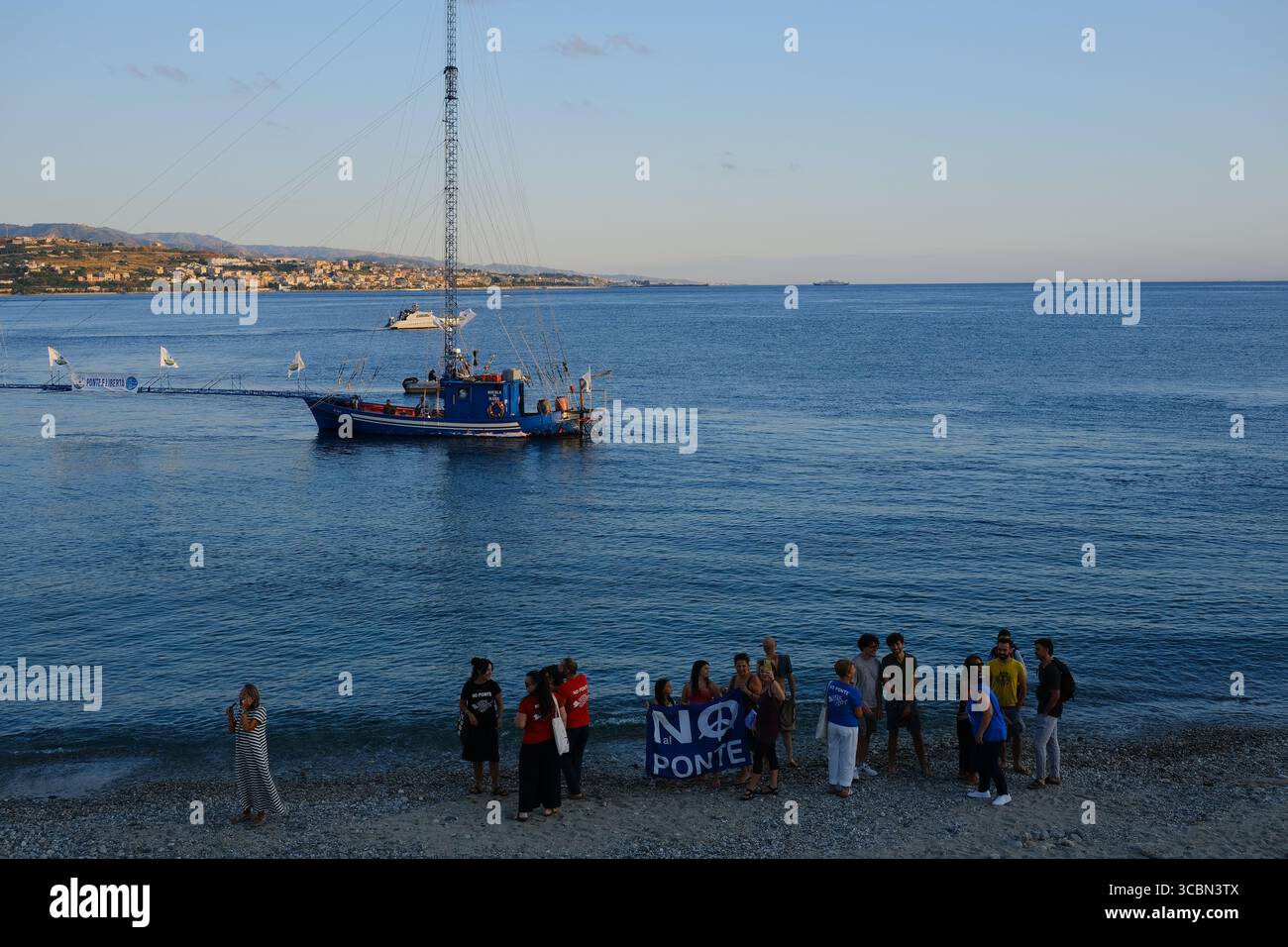 Messine, Sicile, Italie – août 2025. Le vice-premier ministre et ministre de l’infrastructure, Matteo Salvini, assiste à un déjeuner au restaurant Grecale pour célébrer l’approbation par le CIPESS du projet controversé du pont du détroit de Messine. Sur la plage voisine, les baigneurs partagent le sable avec les manifestants tenant un drapeau « No Ponte » (No Bridge) contre la construction. Dans l'eau, un bateau de pêche traditionnel de Messine appelé felouca, utilisé pour la chasse à l'espadon, est décoré de drapeaux italiens de la Lega Nord de droite. Villa San Giovanni en Calabre est visible en arrière-plan. Banque D'Images