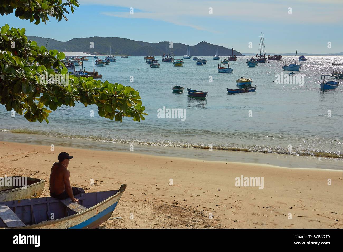 Scène de plage tropicale avec un homme assis sur un bateau en mer avec des bateaux de pêche et feuillage tropical à Buzios, Brésil Banque D'Images