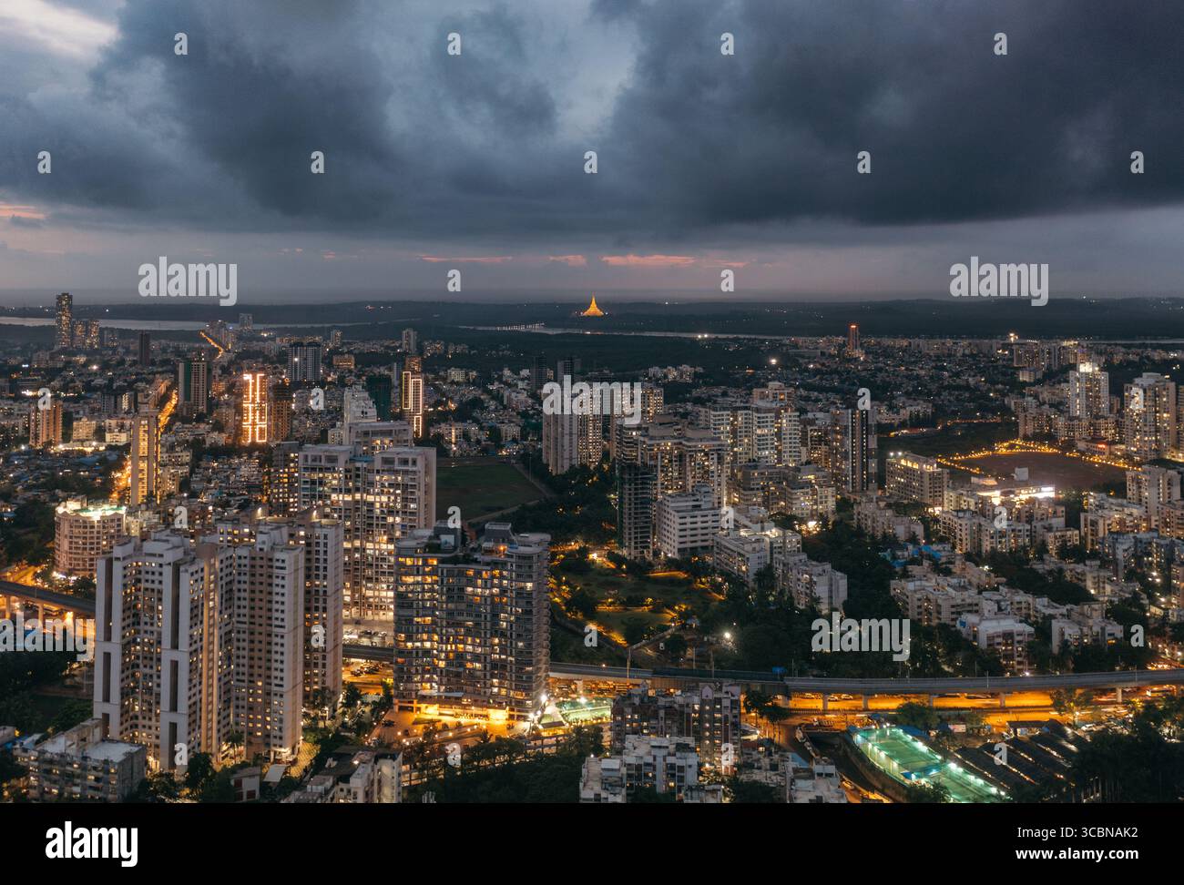 Vue aérienne du paysage urbain baigné dans la lueur chaude des lampadaires contre le ciel crépusculaire, avec la silhouette lointaine de la Pagode Global Vipassana, Mumbai, Maharashtra, Inde. Banque D'Images