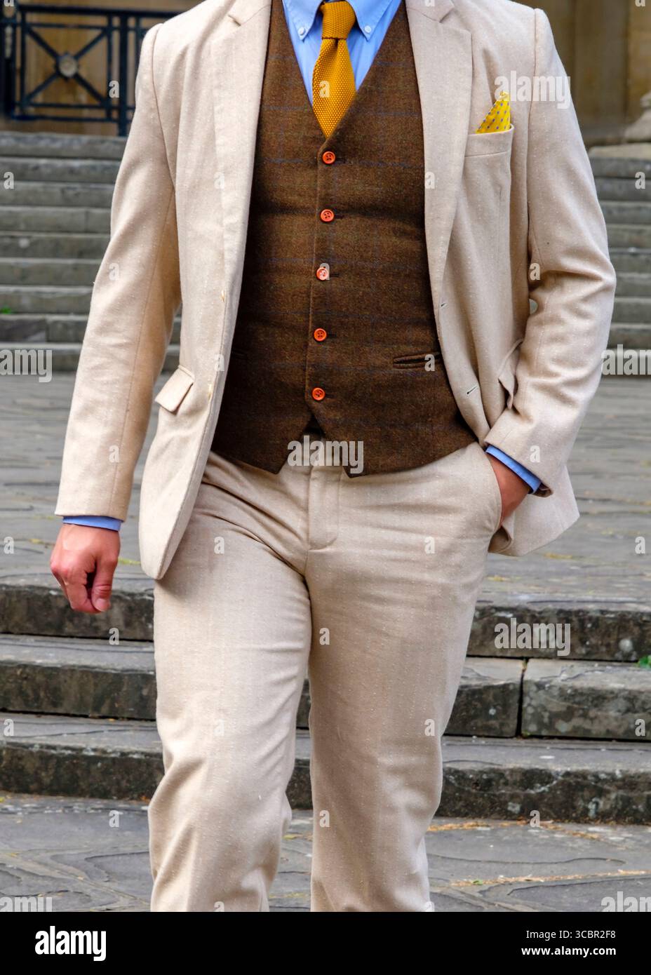 Un homme dans un costume élégant se tient sur un ensemble d'escaliers. Il porte un costume de couleur claire avec un gilet marron, une chemise bleue et une cravate jaune. Un pock jaune Banque D'Images