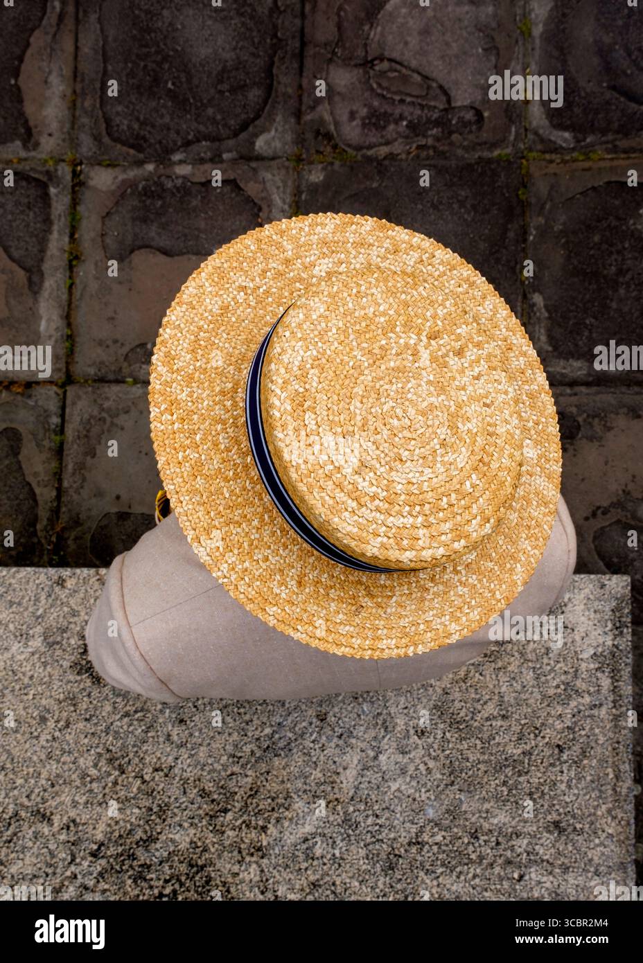 Une photo au-dessus de la tête d'une personne portant un chapeau de paille avec un ruban bleu et blanc, assise sur une marche de pierre. Banque D'Images