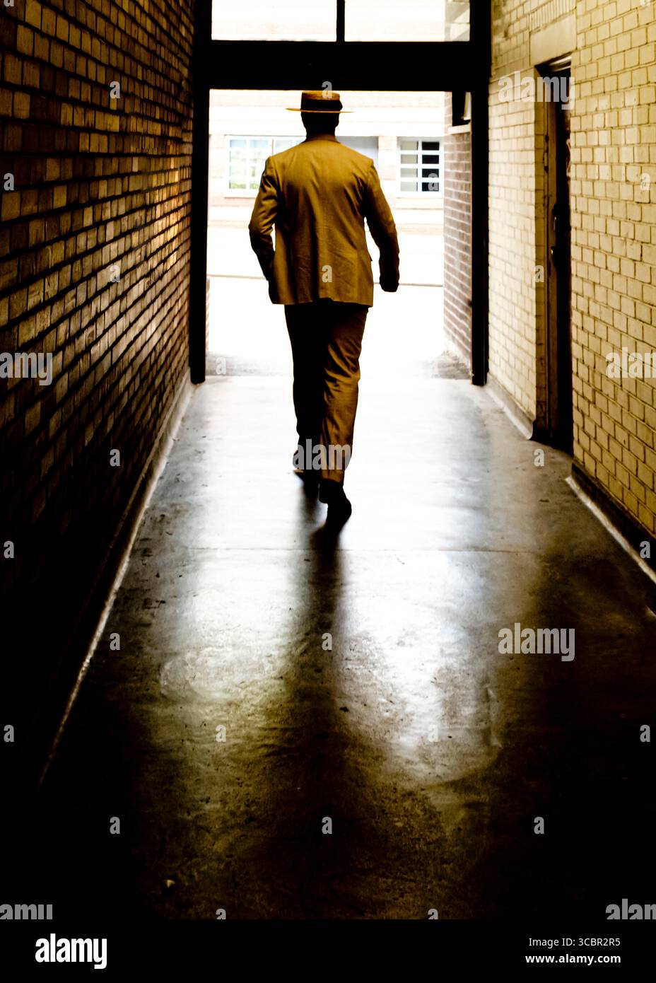 Un homme en costume et chapeau marche dans un couloir sombre vers une porte lumineuse. Banque D'Images