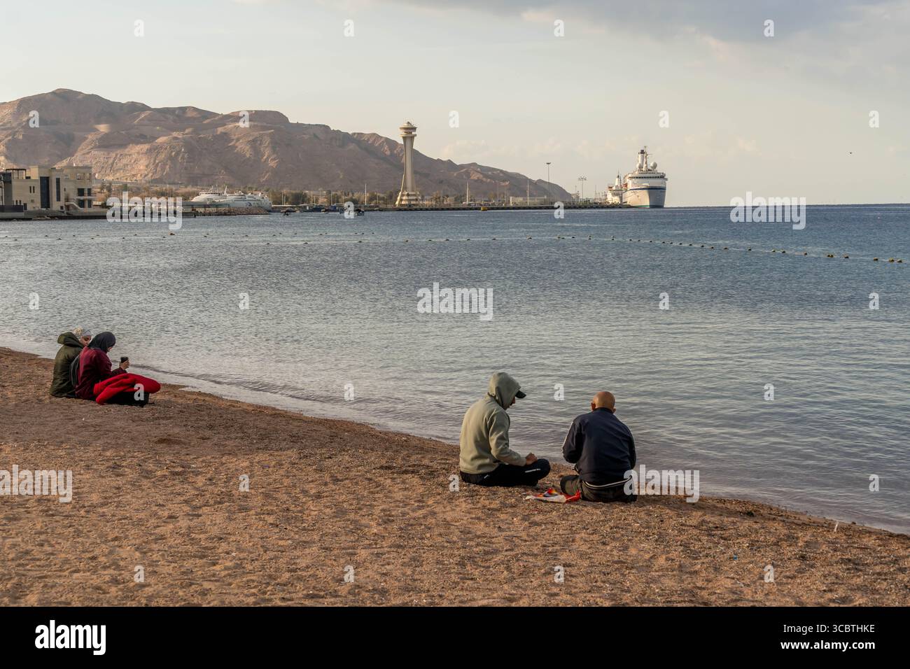 Les habitants jordaniens sont assis sur la rive de la mer Rouge avec le port d'Aqaba et les montagnes en arrière-plan, profitant d'un moment de calme au bord de l'eau. Banque D'Images