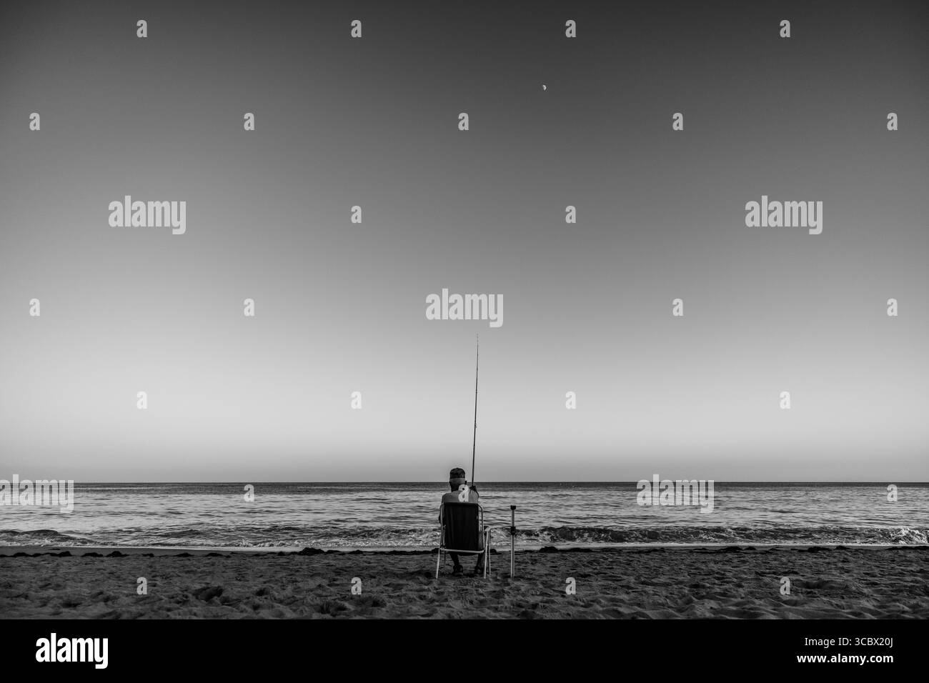 Algarve, Portugal – 2025.07.03 : un homme est assis sur une chaise avec une canne à pêche, face à la mer calme sous un ciel clair du soir, alors que la lune se lève au-dessus du h. Banque D'Images