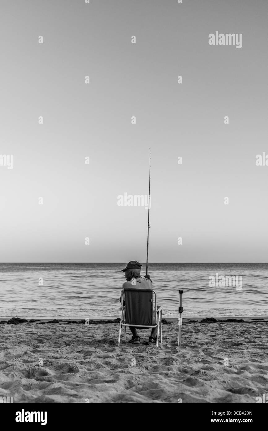 Algarve, Portugal – 2025.07.03 : un homme est assis sur une chaise avec une canne à pêche, face à la mer calme sous un ciel clair du soir, alors que la lune se lève au-dessus du h. Banque D'Images