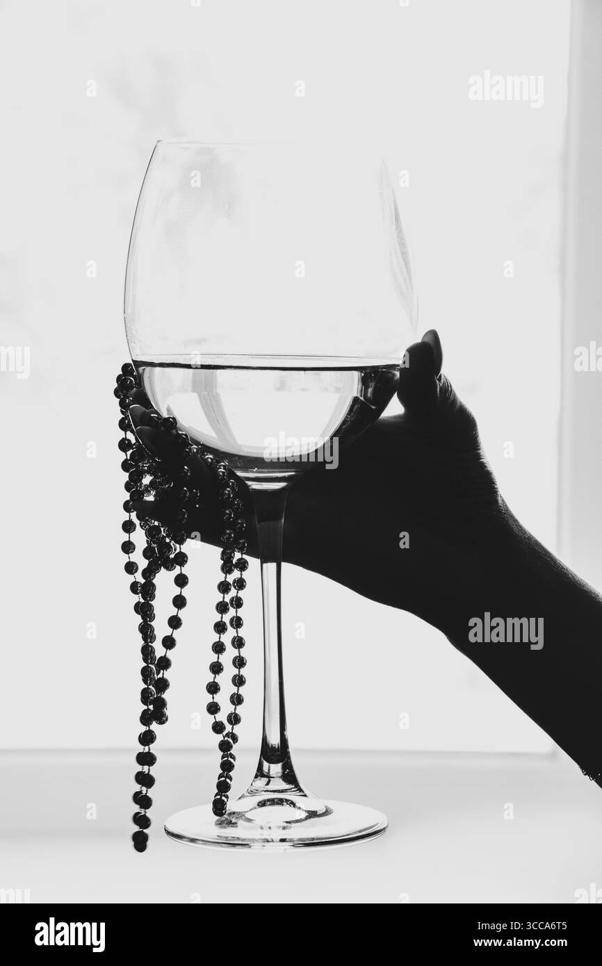 Main de femme avec manucure tenant un verre de vin blanc et des perles par la fenêtre dans une pièce lumineuse Banque D'Images