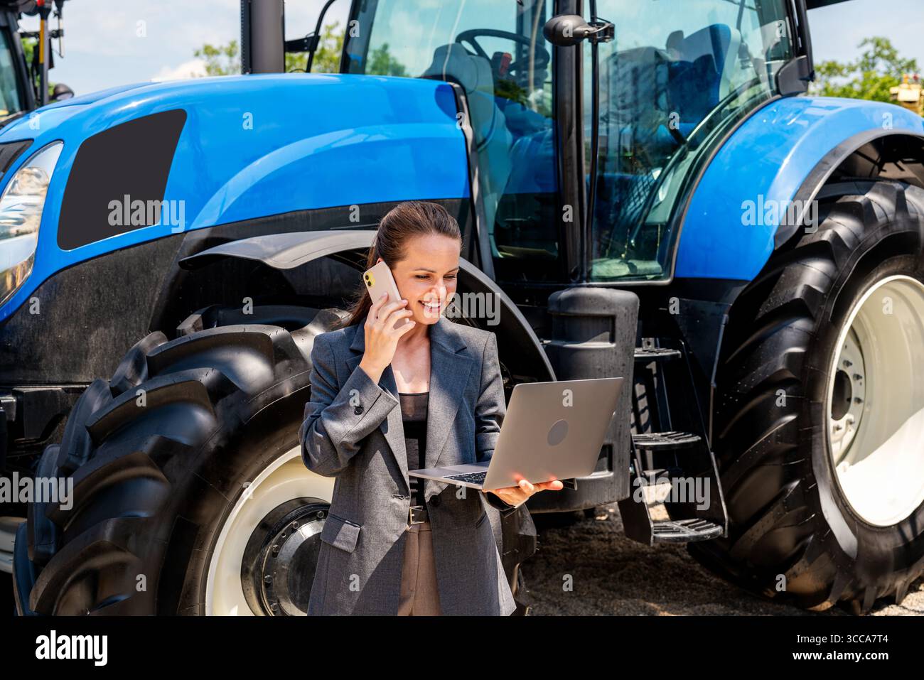 Femme d'affaires avec ordinateur portable et téléphone portable debout devant le nouveau tracteur à la concession de matériel agricole Banque D'Images