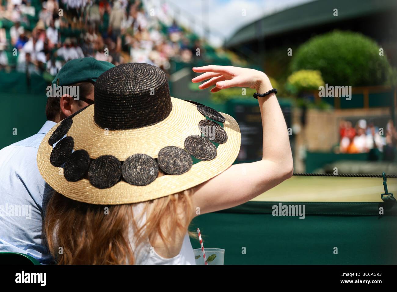 Jeune femme portant un grand chapeau de paille assis sur l'un des courts extérieurs aux championnats de Wimbledon 2025, Londres, Angleterre, Royaume-Uni. Banque D'Images