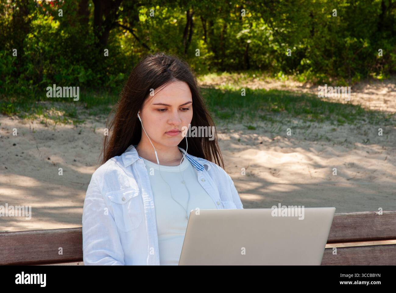 Jeune femme avec de longs cheveux bruns travaillant sur un ordinateur portable avec des écouteurs tout en étant assise sur un banc en bois dans un parc de sable ensoleillé, symbolisant le travail à distance, fr Banque D'Images